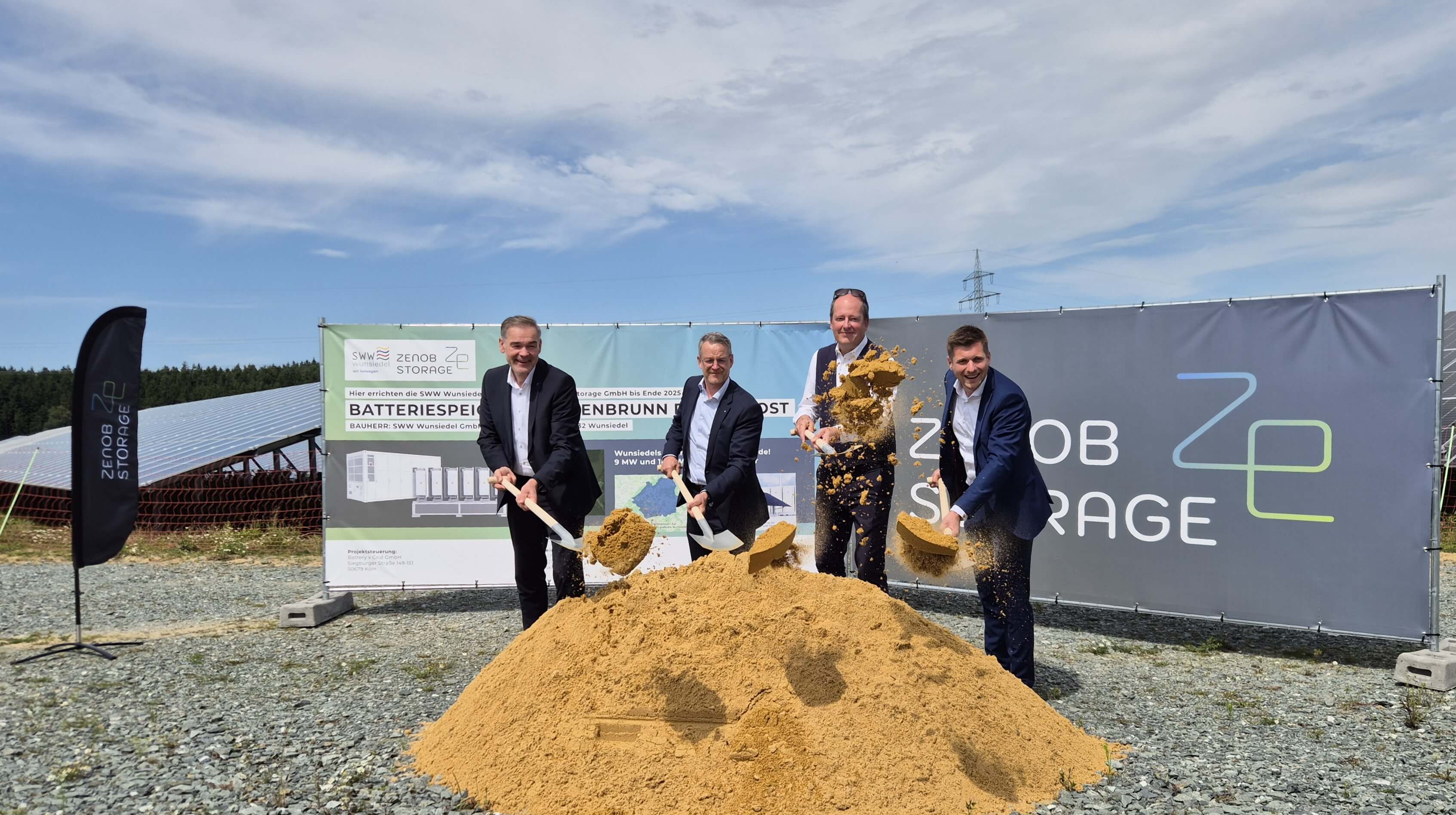 Peter Berek, Marco Krasser, Winfried Karl und Nikolas Lahovnik stechen jeweils mit einem Spaten einen Erdhaufen an. Im Hintergrund sind der Solarpark Holenbrunn und Bauzaunbanner mit Infos zu ZENOB Storage zu sehen.