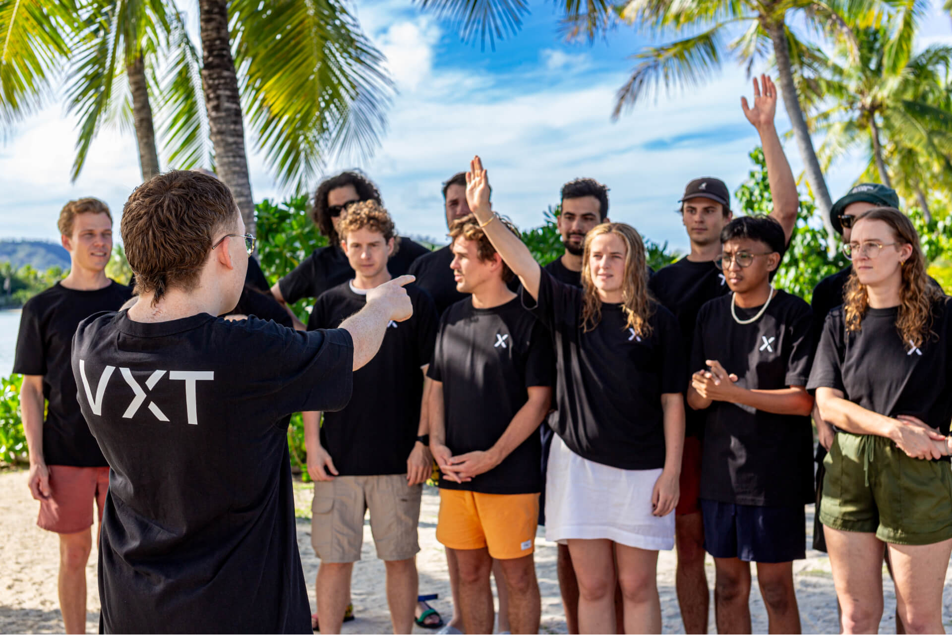 Group of young people wearing matching black shirts outdoors, with one person raising their hand and another pointing.