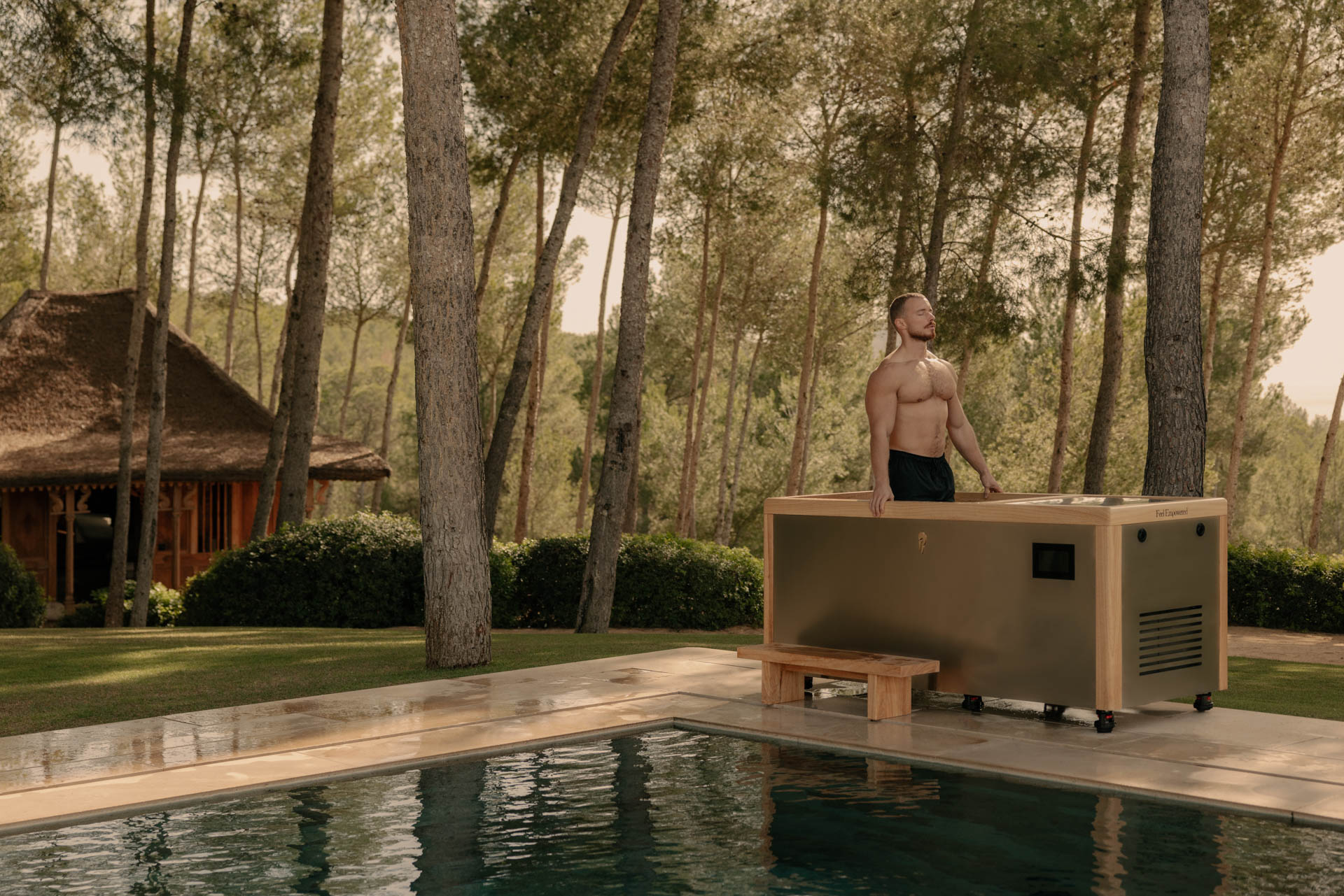 Uomo in piedi in una vasca di immersione a freddo all’aperto vicino a una piscina, circondato da pini, in un contesto naturale e premium.