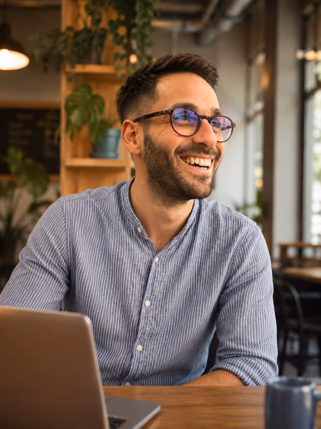 Une personne souriante portant des lunettes et une chemise bleu clair à fines rayures est assise à une table avec un ordinateur portable dans un café lumineux. Des plantes et un éclairage chaleureux créent une ambiance accueillante en arrière-plan.