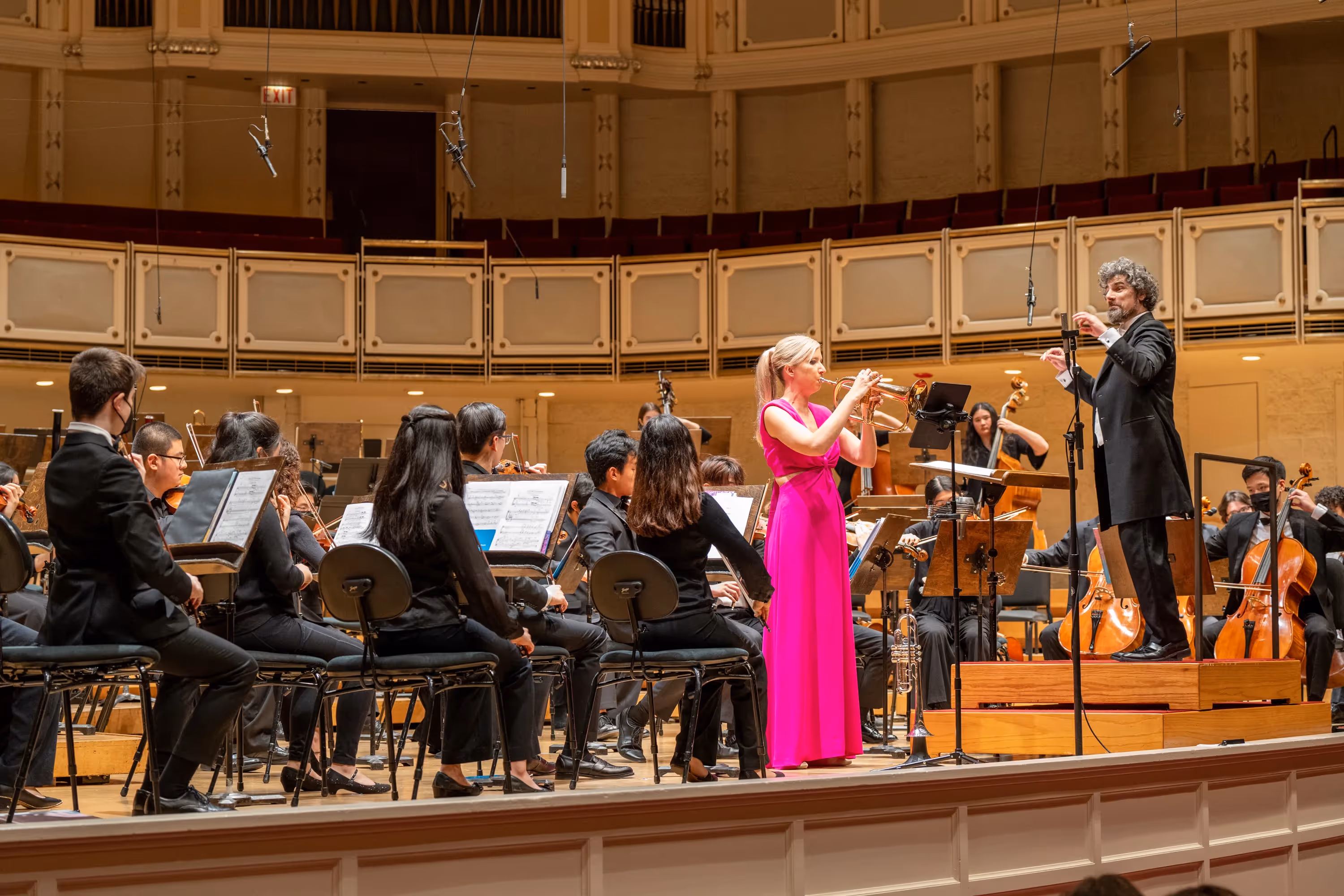 Mary Elizabeth Bowden performing the world premiere of Clarice Assad's Bohemian Queen: Concerto for Trumpet and String Orchestra with the Chicago Youth Symphony Orchestra. Bowden is at front of the stage wearing a hot pink dress. Conductor Allen Tinkham is in a suit, and members of the orchestra are wearing all black.