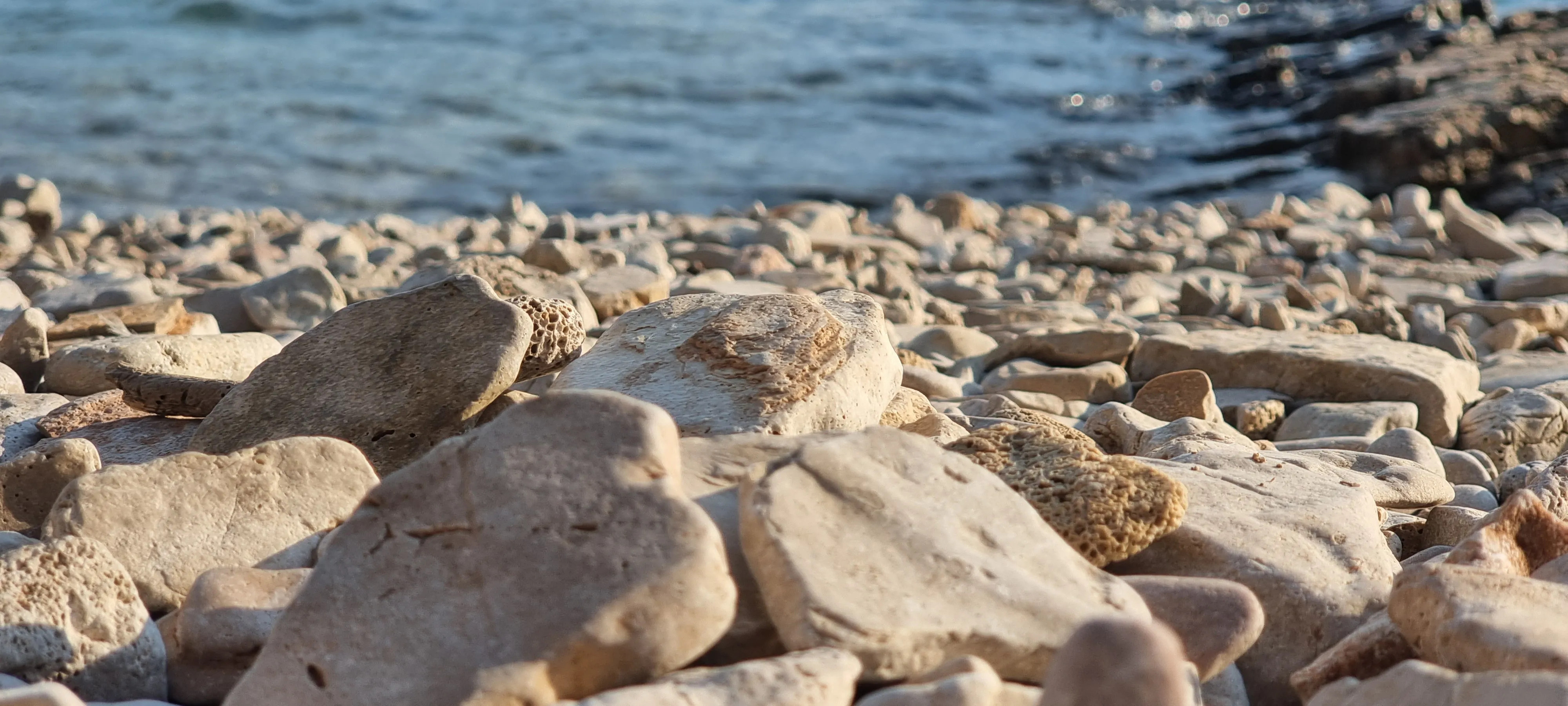 close up of white pebbles on a pebble beach with shore and water in background - the gymsy traveller