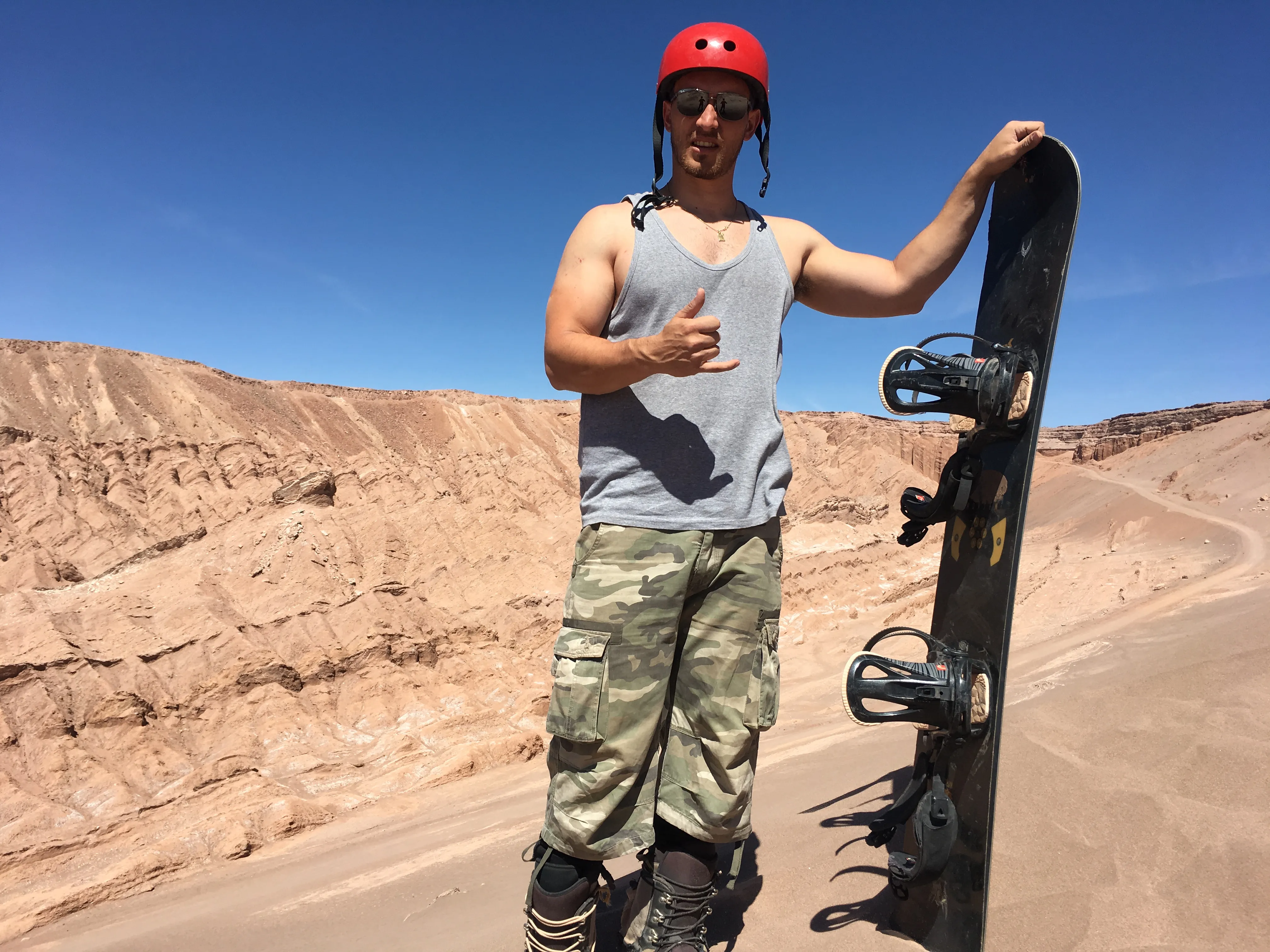 the gymsy traveller posing at the top of a sand dune/hill with a snowboard held beside him, blue skies and lots of desert and sand surroundings in the atacama desert in chile