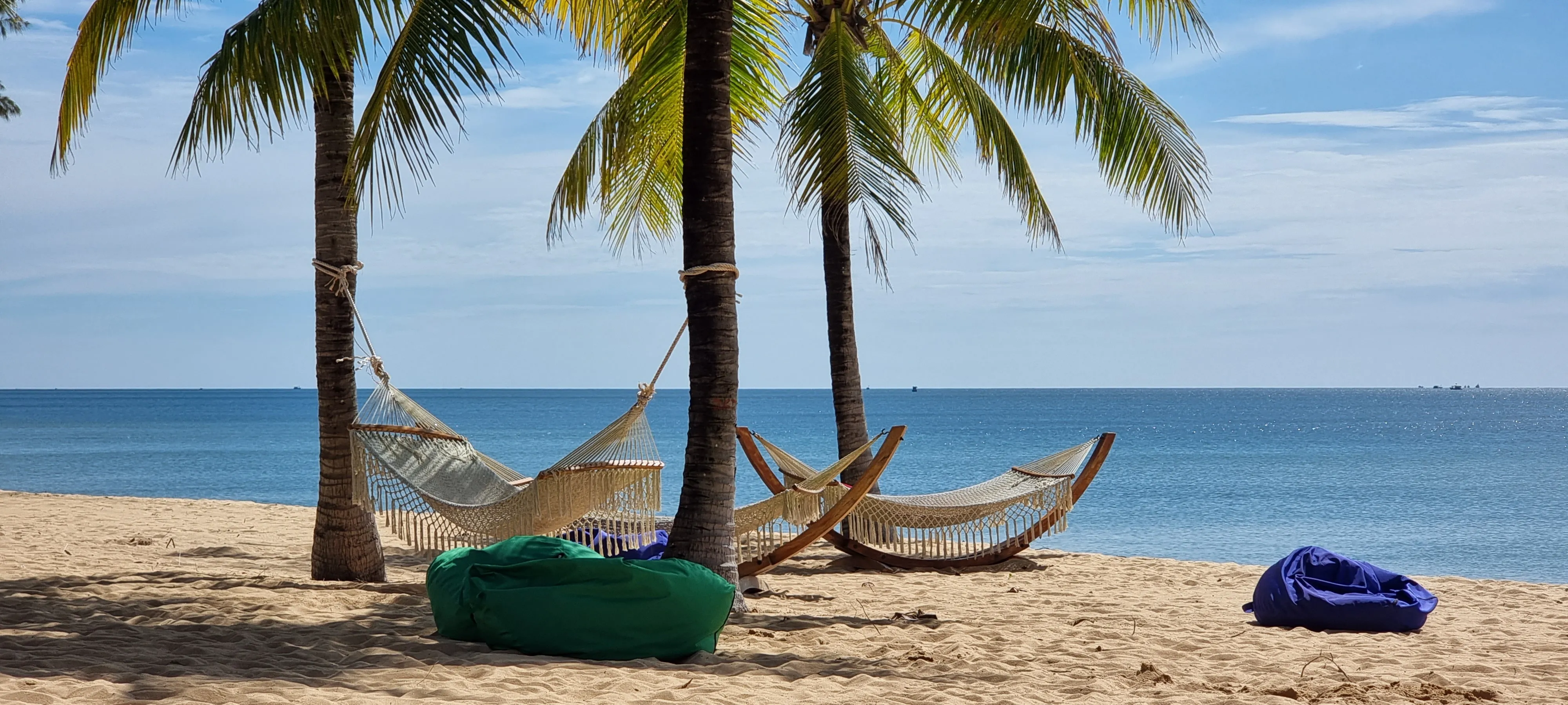 a serene image overlooking a beach in phu quoc island vietnam. three hammocks in view with some beanbags scattered on the golden sand. palm trees holding up the hammocks, blue sea in background and blue skies above. thegymsytraveller 