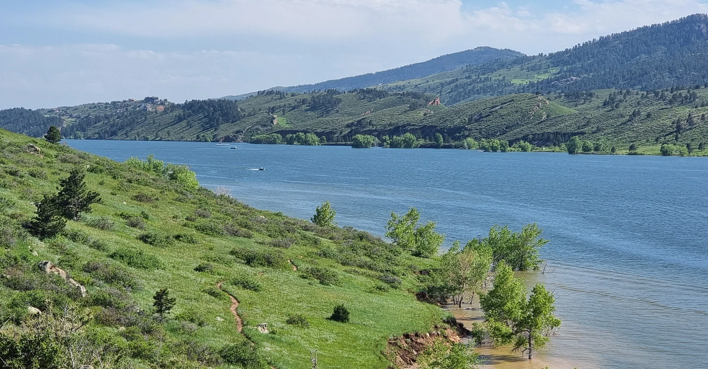 horsetooth reservoir in colorado, picture from a steep hill overlooking the lake and lush hills in the background. one or two speedboats in the lake, great hiking trails can be seen