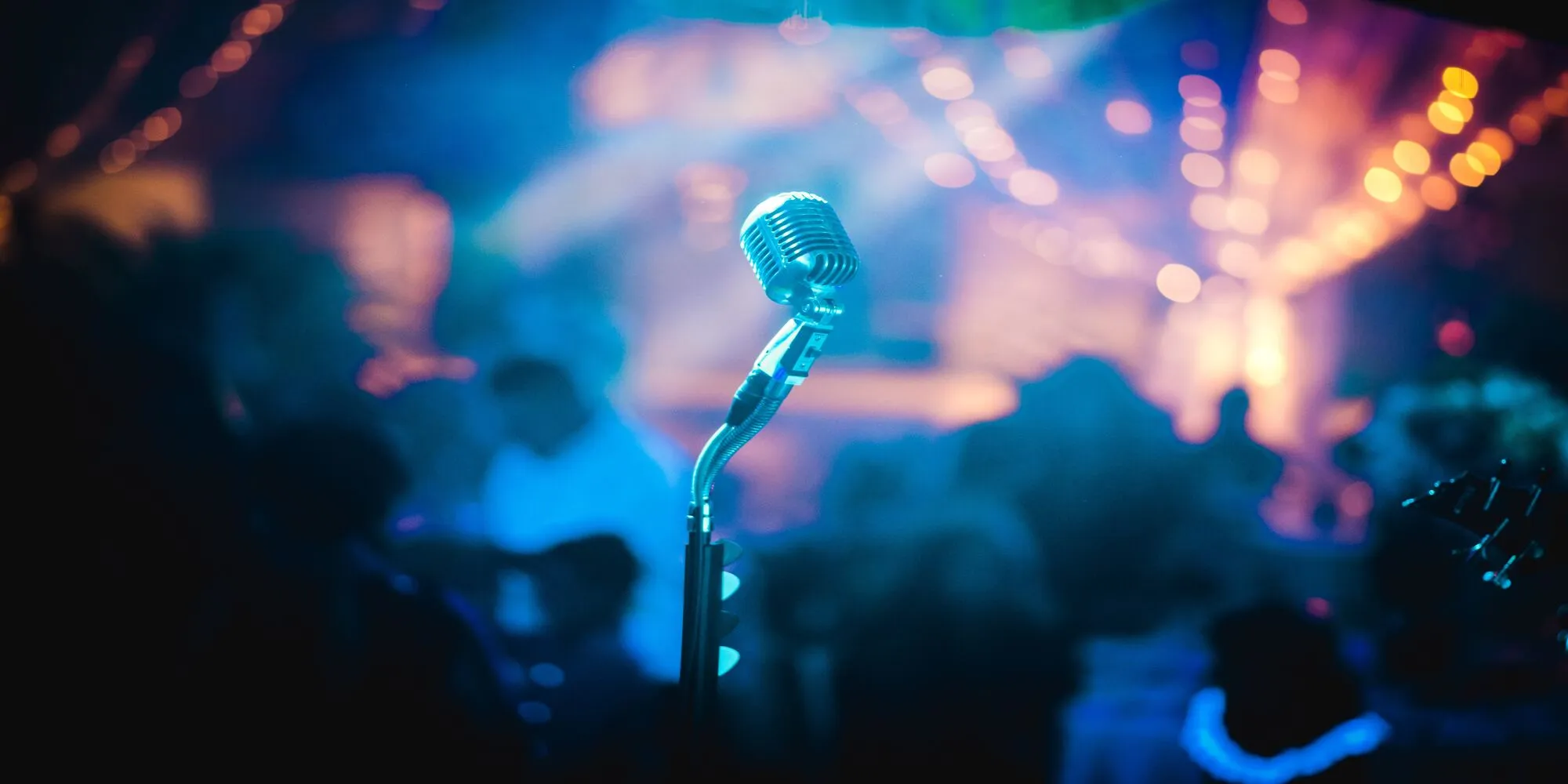 an image of an old microphone in the middle of a room and stage, background is blurred but bright colours and a crowd sit surrounding the mictrphone