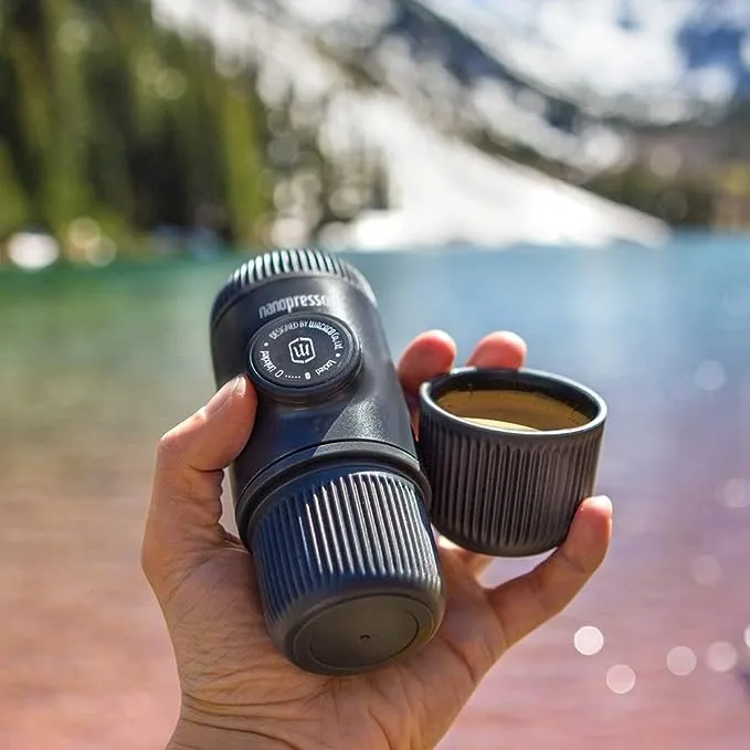 an image of a small coffee maker being held in a persons hand. the background is blurred but you can still make out a mountain scene with a lake