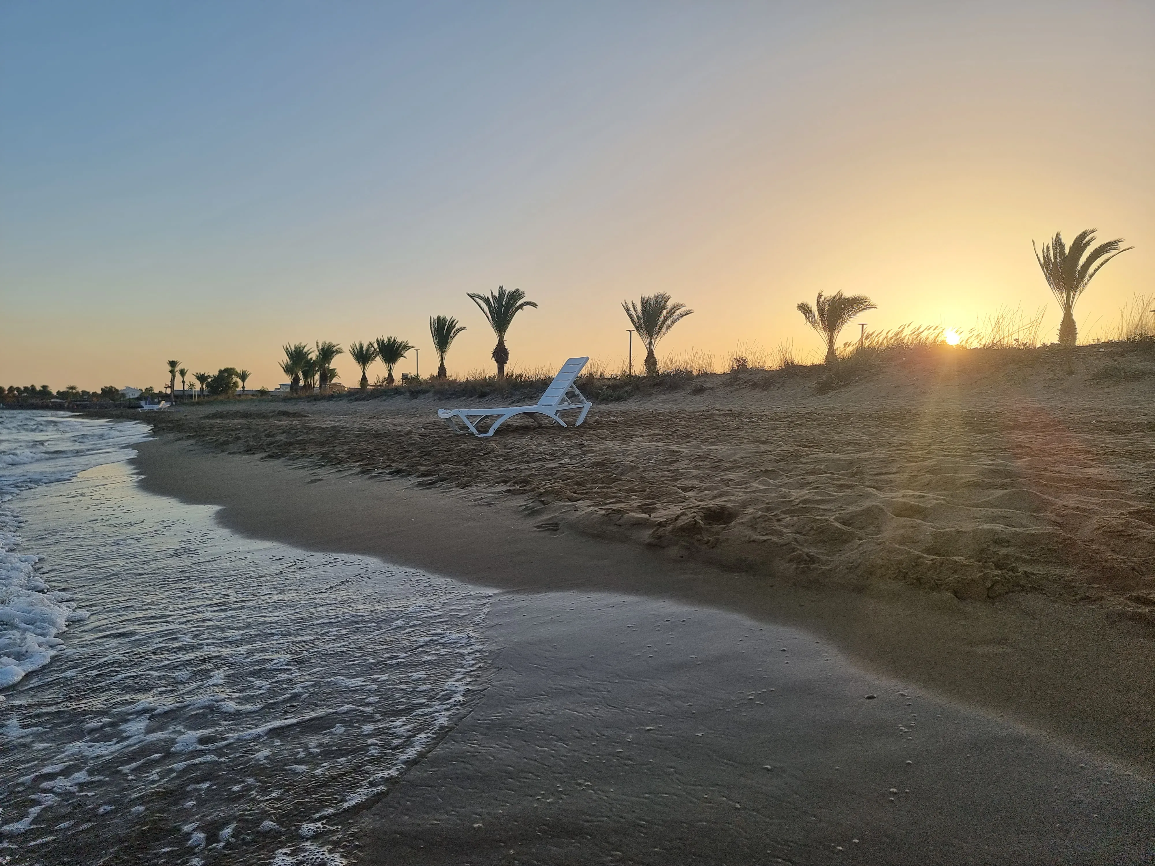 an image of a beach in famagusta cyprus in november, a single beach lounge is in the middle of the beach with palm trees in he background. sunsetting in the distance and clear skies above
