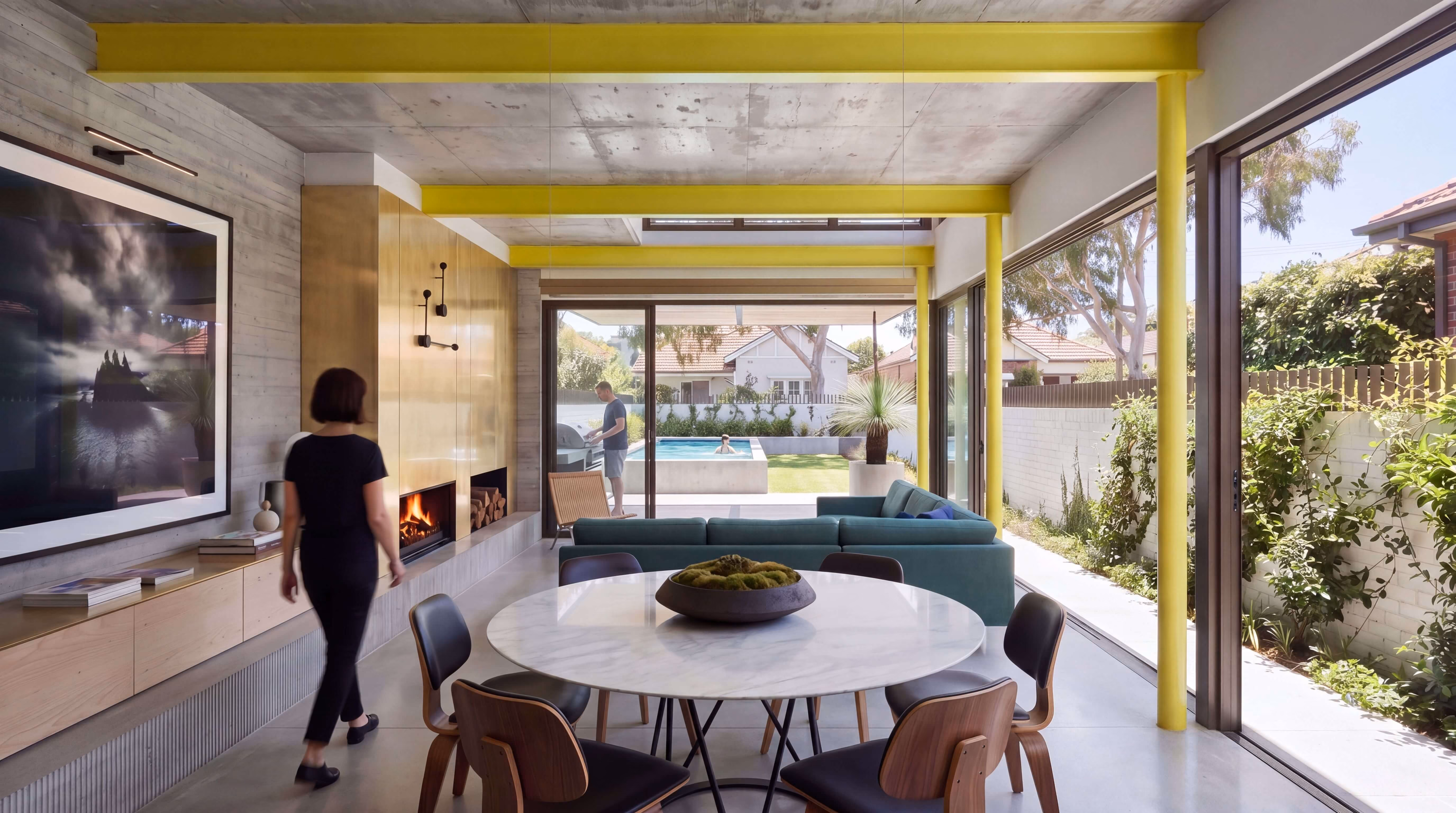 Modern living and dining room with large floor-to-ceiling windows overlooking a backyard with pool, featuring yellow ceiling beams, a round marble dining table with chairs, teal sectional sofa, wood-paneled wall with fireplace, and a woman walking inside.