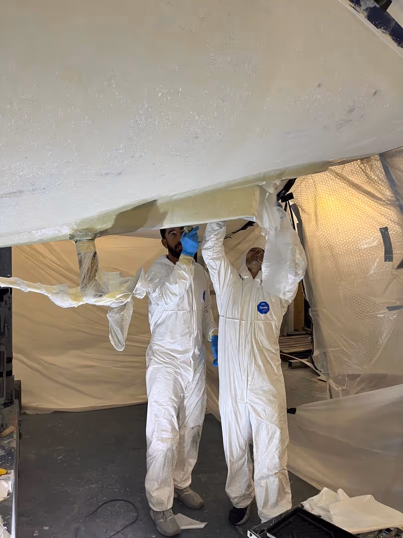 Two people in white protective suits work under a large, white fiberglass object.
