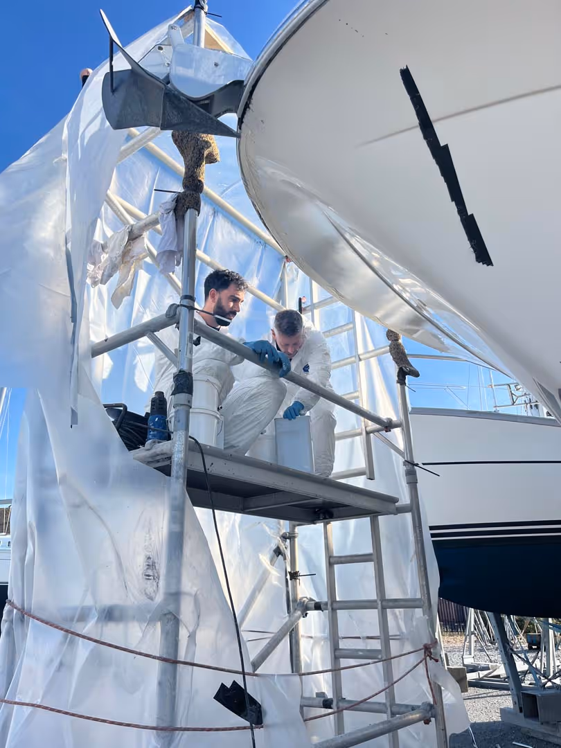 Two people in protective suits work on scaffolding under a covered boat.