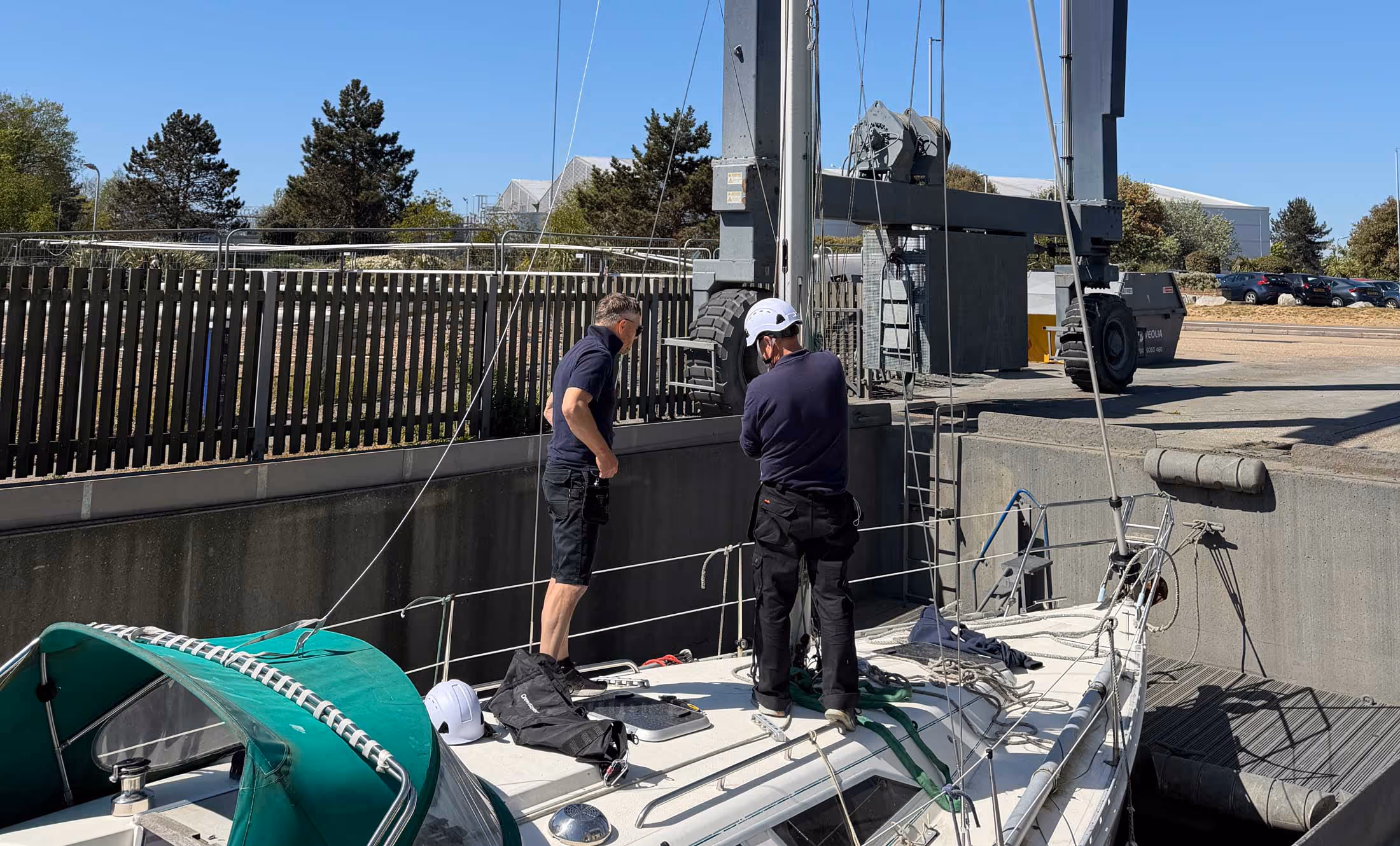 Two men working on a sailboat in a dock area, surrounded by equipment and fencing.