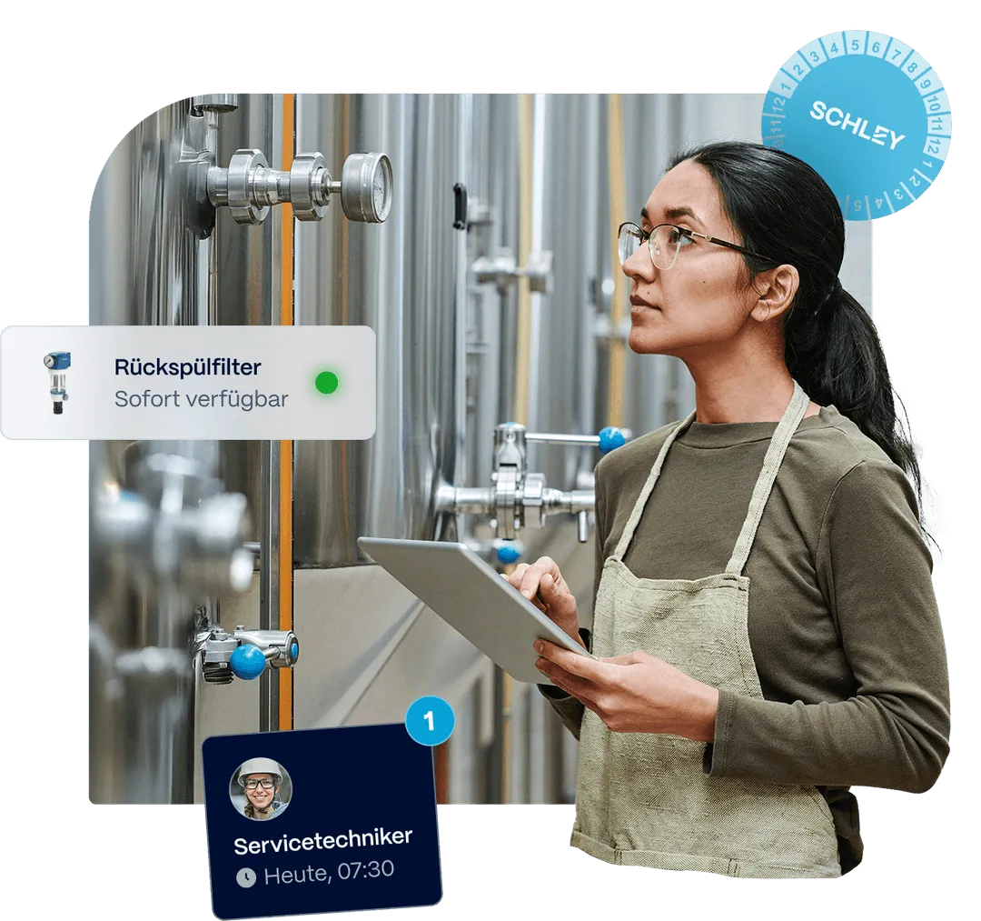 A woman in an apron inspects large metal tanks, holding a tablet in a workspace.