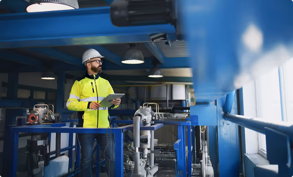 A worker in a bright safety jacket and helmet holds a tablet in a factory setting.
