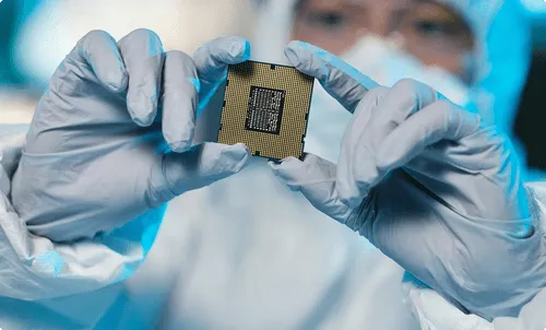 A person in protective gear holds a microchip with both hands, focusing on its surface details.