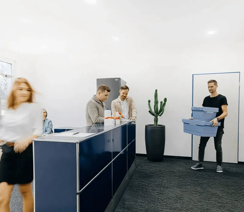 A person walks past two others at a desk while another carries stacked plastic boxes.
