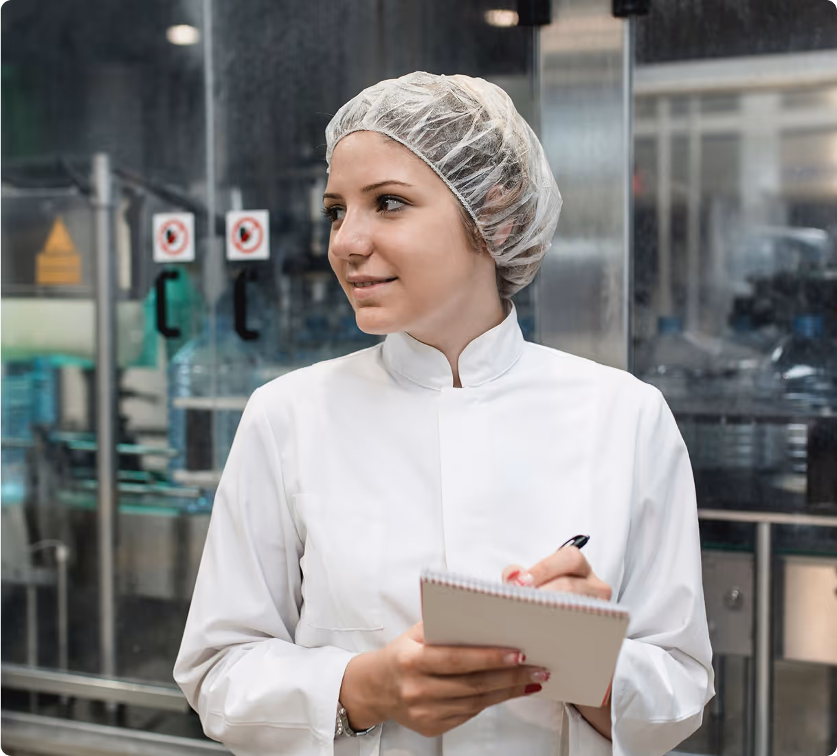 Woman in a lab coat and hairnet taking notes in an industrial setting.