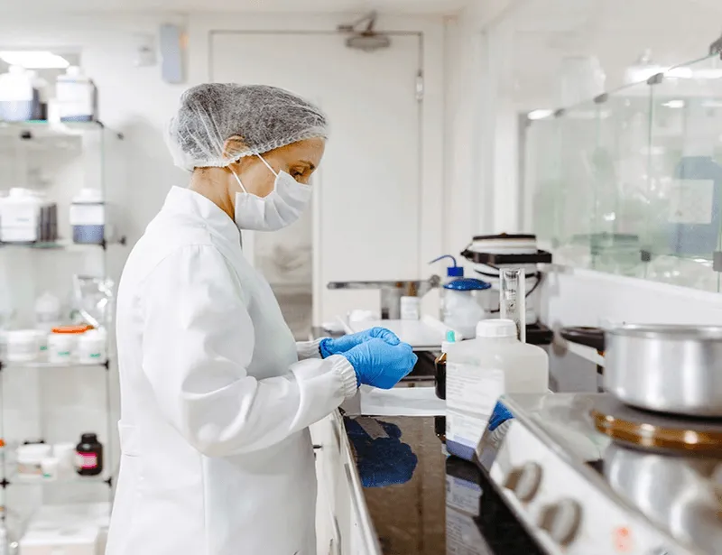 A person in a lab coat and mask works in a laboratory, examining a sample.