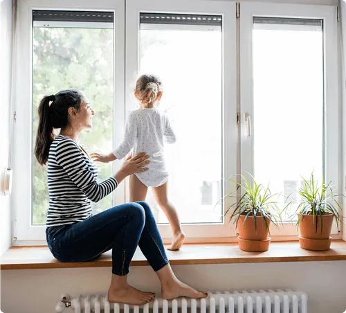 Woman sitting on a windowsill helps a toddler standing and looking outside.