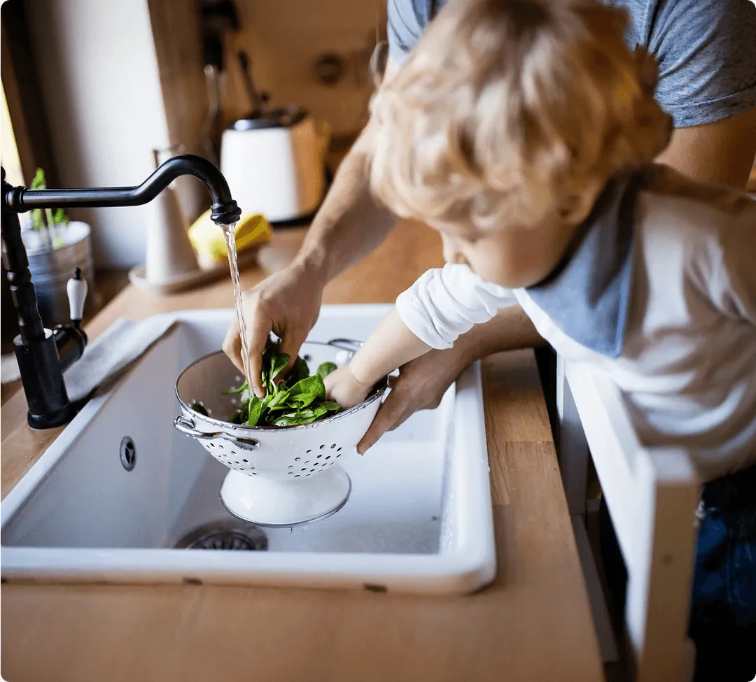 Child helps adult wash spinach in a kitchen sink using a colander.