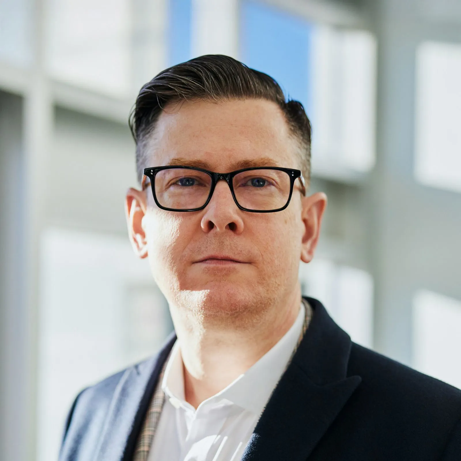 A person wearing glasses looks directly at the camera, standing in a sunlit room.
