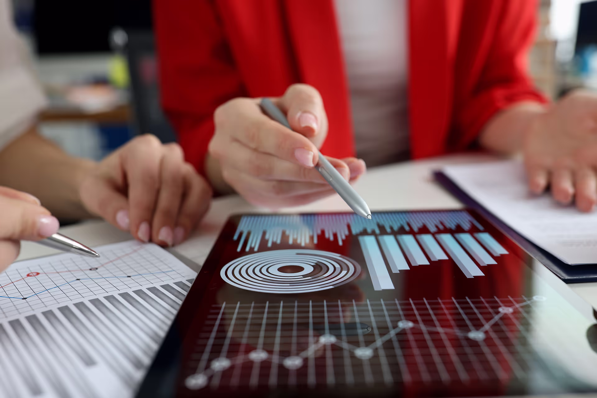 Hands holding pens pointing at tablet displaying various digital data charts and graphs with printed documents on table.