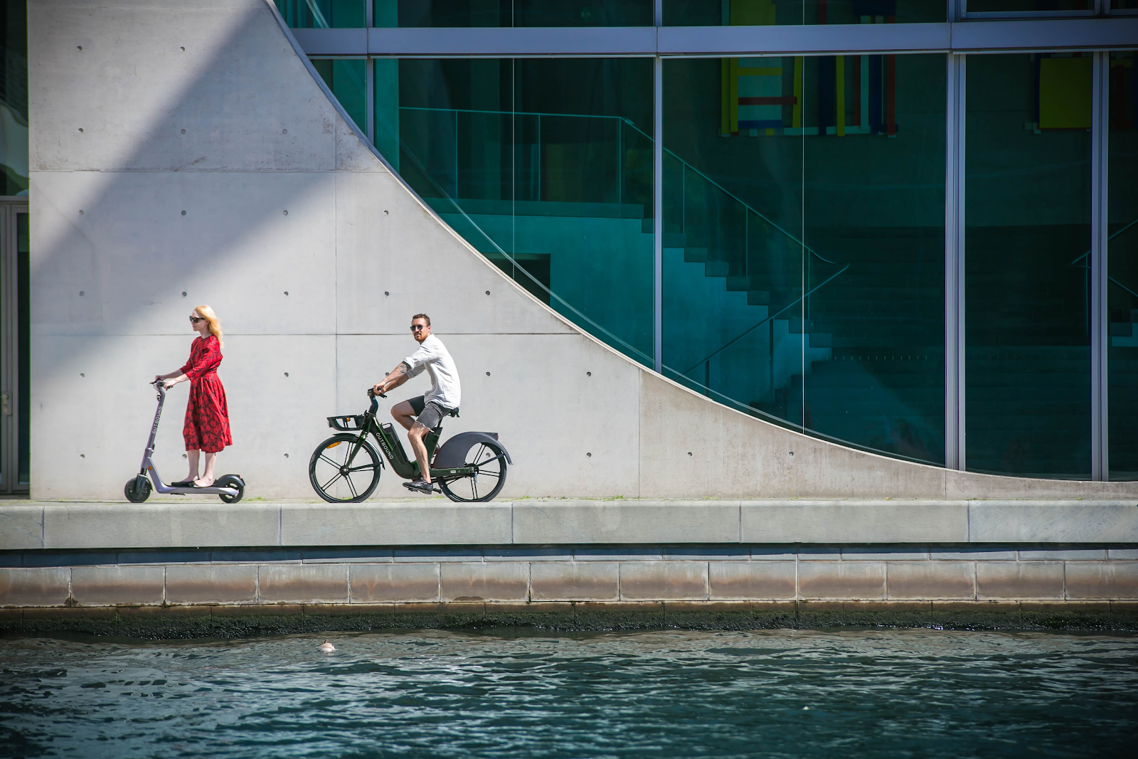A young woman enjoys riding an e-scooter and a young man enjoys riding an e-bike outside a modern property