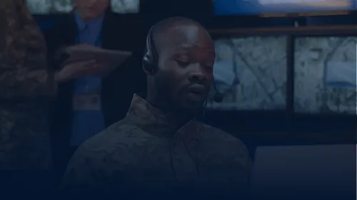 Hombre con uniforme militar y auriculares hablando en un centro de mando con varios monitores al fondo.