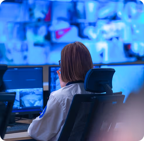 Person with short hair wearing glasses and a white jacket monitoring multiple security camera feeds on screens.