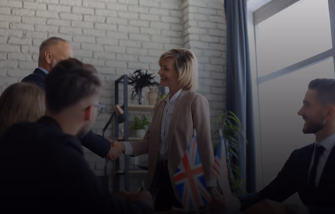 Two businesspeople shaking hands in an office while colleagues watch, with British and American flags on the table.