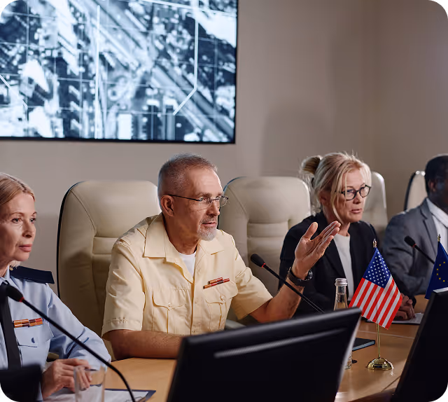 Des responsables militaires et civils assis autour d'une table de conférence, sur laquelle sont posés des drapeaux américains et européens ainsi que des écrans, dans une salle de briefing.