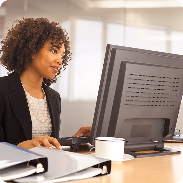 Une femme aux cheveux bouclés travaillant sur un ordinateur de bureau, assise à son bureau, avec une tasse de café et des classeurs.