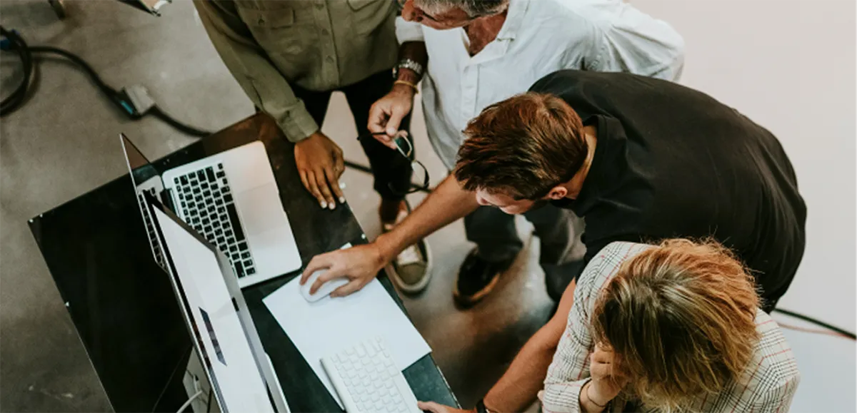 Four colleagues collaborating around a desk with a laptop and computer monitor, viewed from above.