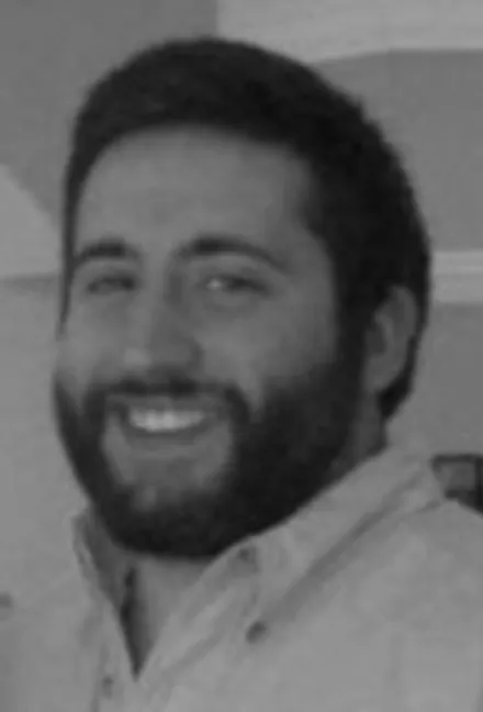 Smiling man with dark hair and beard wearing a light-colored collared shirt.