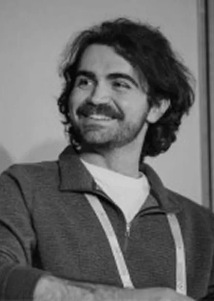 Smiling man with dark curly hair and a beard wearing a zip-up jacket over a light-colored shirt.