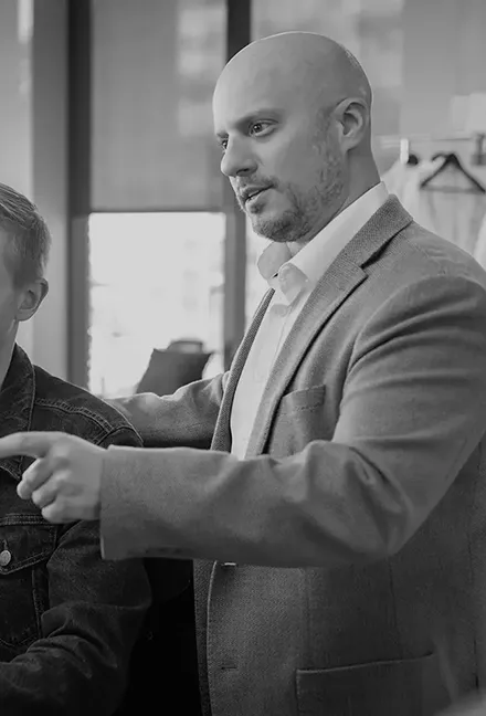 Bald man in a suit jacket pointing forward while engaged in conversation with another person.