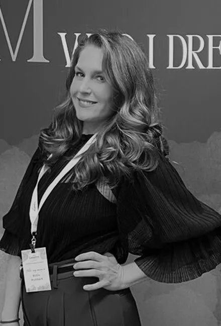 Smiling woman with wavy hair wearing a pleated blouse and conference badge posing with hand on hip.