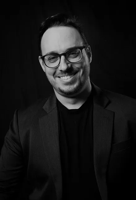 Smiling man wearing glasses and a dark blazer against a black background.