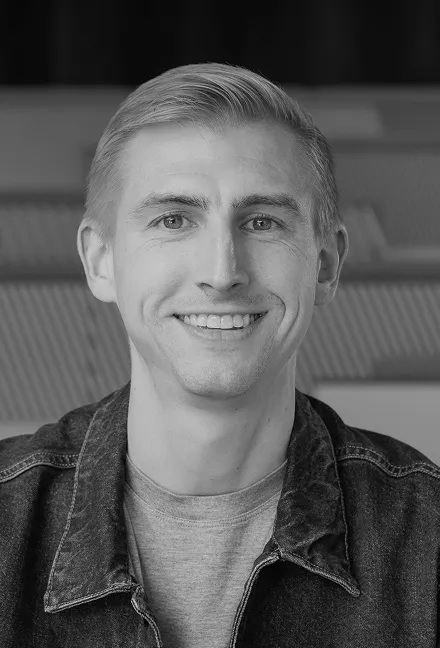 Smiling young man with short, light-colored hair wearing a denim jacket and gray shirt, sitting indoors.