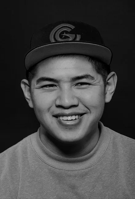 Black and white portrait of a smiling young man wearing a cap and a crewneck sweatshirt against a dark background.