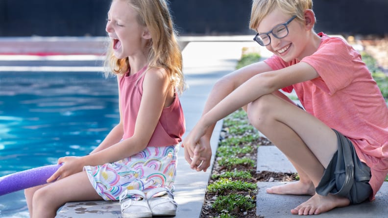 A girl and a boy, laughing, sitting next to a swimming pool