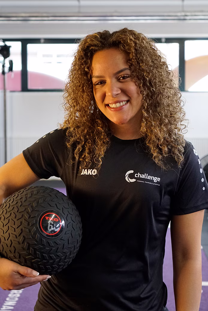 Laura Vinken - Smiling woman with curly hair holding a 6 kg black textured medicine ball in a gym setting.