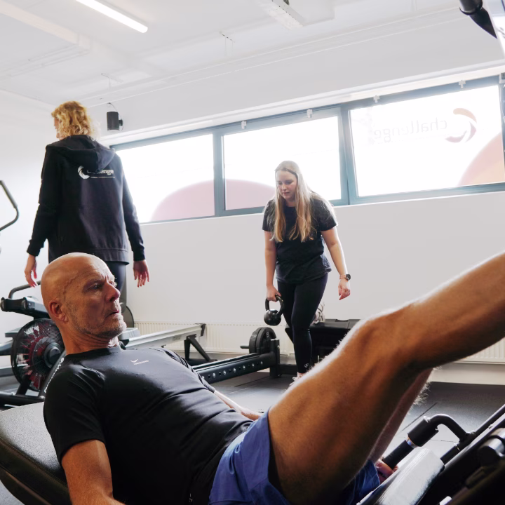 Older man exercising on a leg press machine in a gym with two women in the background holding kettlebells and walking.