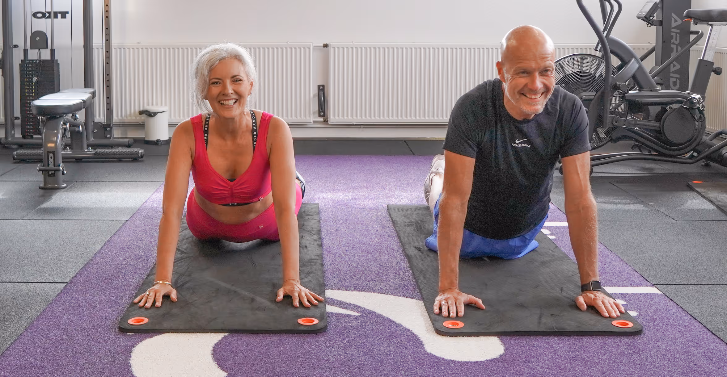 Smiling man and woman doing yoga stretches on mats in a gym setting.