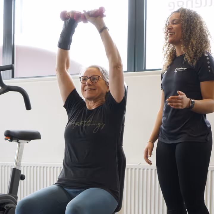 Older woman lifting pink dumbbells overhead while a fitness trainer coaches her in a gym.