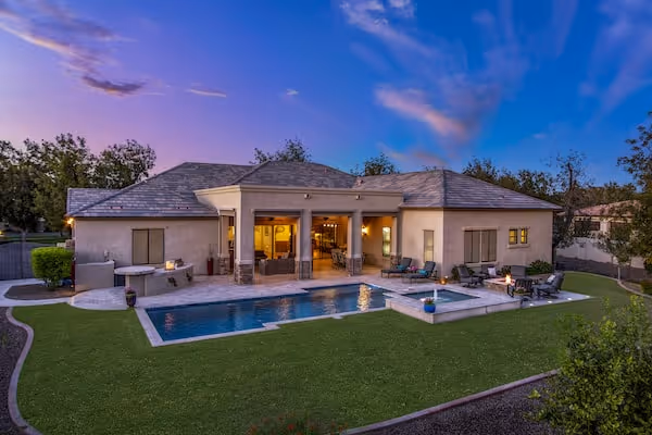 Elegant single-story home in Queen Creek, Arizona, featuring a spacious backyard with a swimming pool, spa, lounge seating, and manicured lawn. The house has a covered patio and large sliding doors opening to a warmly lit interior, with a sunset sky casting soft purple and pink hues overhead.