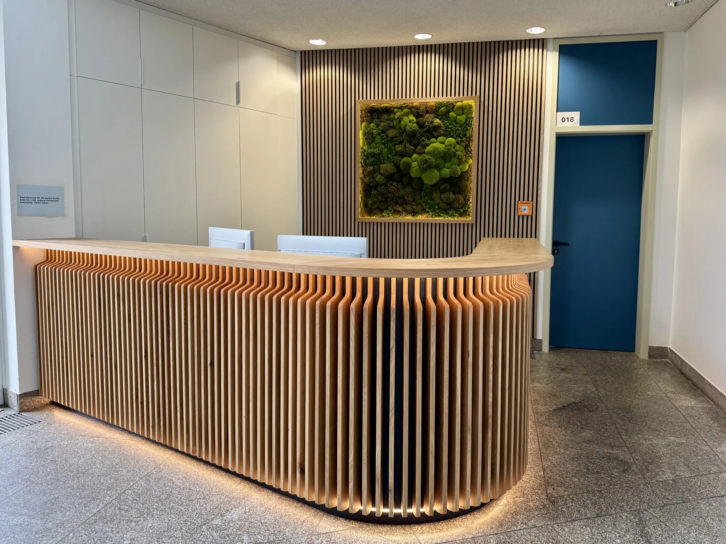 Empfangstresen aus Eiche mit indirekter Beleuchtung und Moosbild im Hintergrund, inklusive Einbauschrank und Wandverkleidung mit Akustikplatten