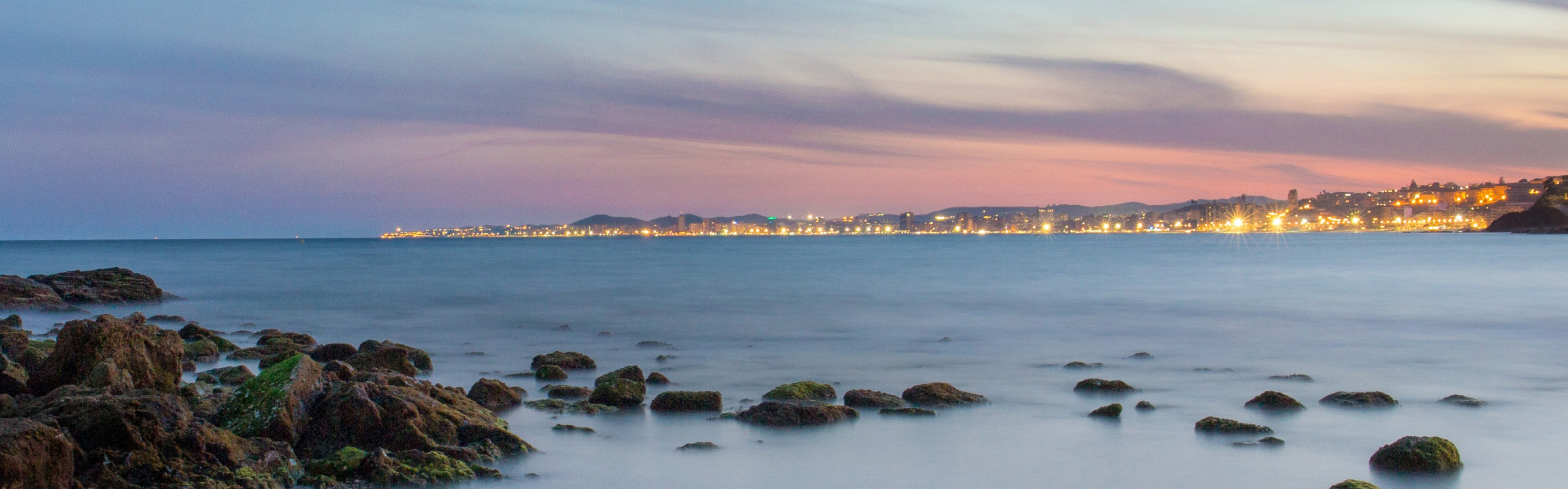 Imagen de la costa de Fuengirola, Málaga en pleno atardecer con las luces de la ciudad al fondo. 