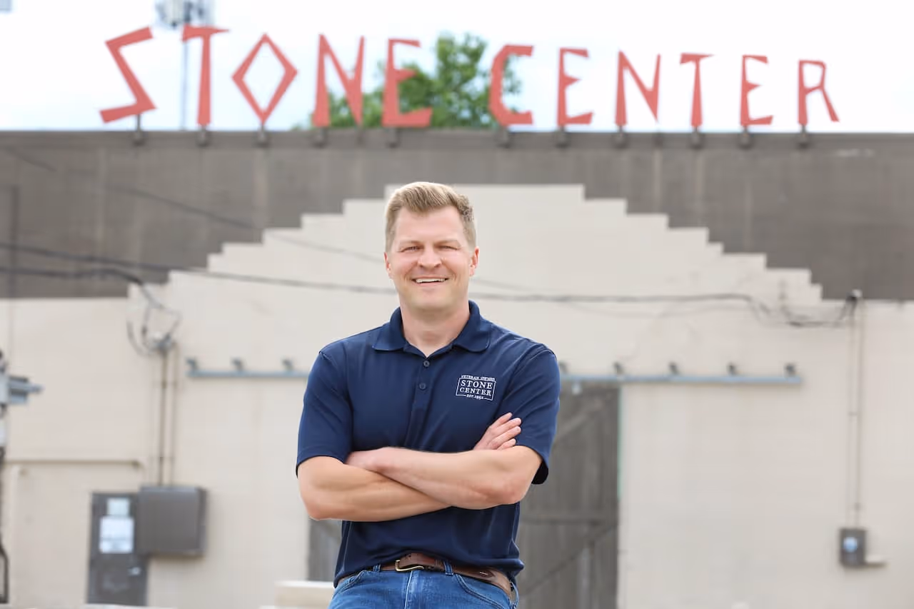 Smiling man in a navy blue Stone Center polo shirt with arms crossed standing in front of a building with a red 'STONE CENTER' sign.