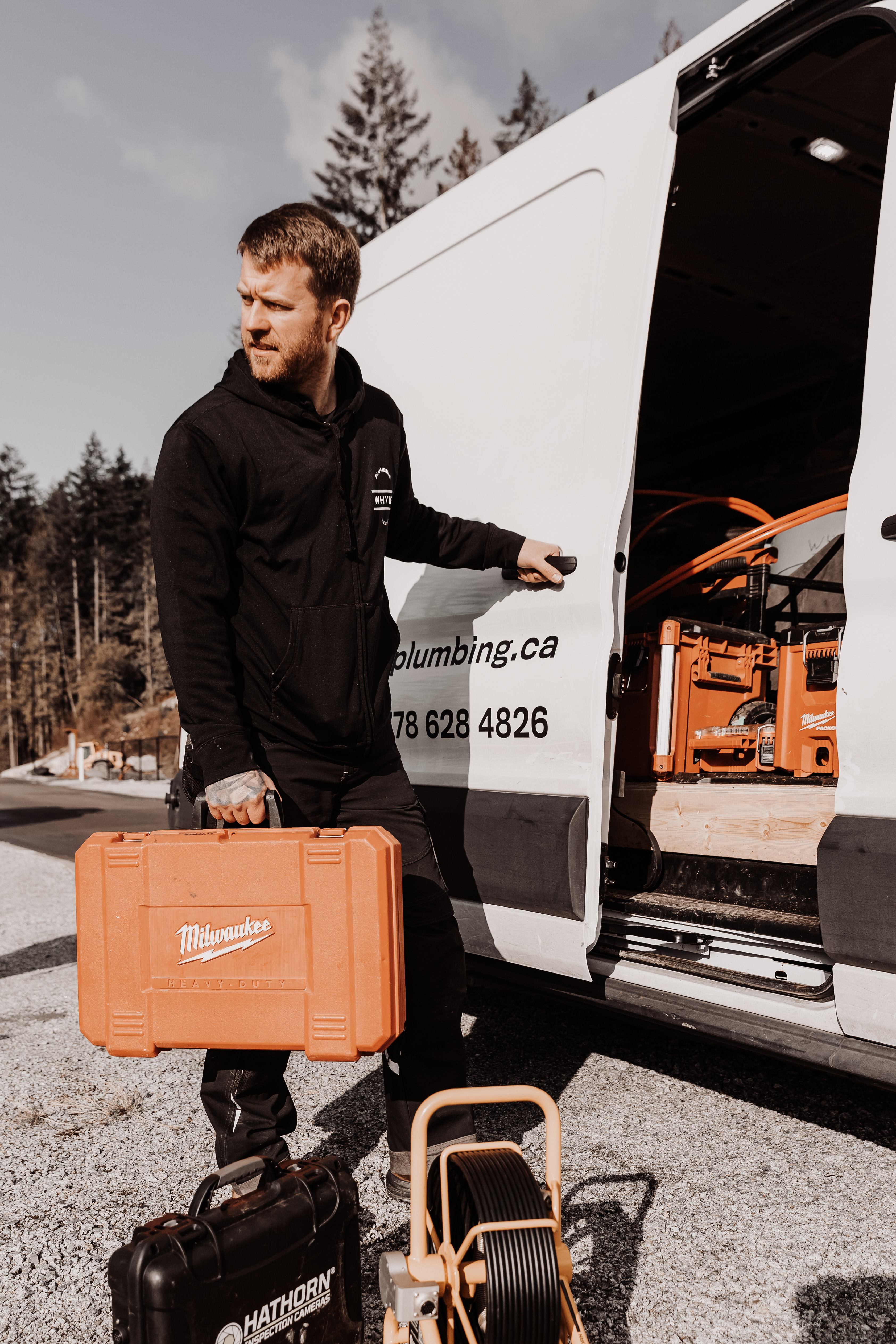 Man in black hoodie holding orange Milwaukee tool case next to open white work van.